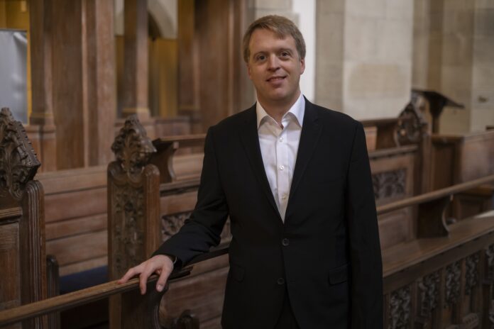 Das Bild zeigt Stefan Donner in dunklem Anzug, der lächelnd in einem Kirchenraum mit hölzernen Sitzbänken steht und in die Kamera blickt.