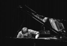 Simon Raab – Solo Piano / Kulturforum New York Schwarzweißfoto eines Pianisten am geöffneten Konzertflügel. Der Musiker beugt sich konzentriert über die Klaviersaiten und greift mit einer Hand in den Flügel, während ein Mikrofon nahe der Saiten positioniert ist. Das Bühnenlicht hebt Hände, Saiten und Mechanik hervor, der Hintergrund bleibt dunkel.