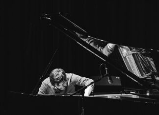 Simon Raab – Solo Piano / Kulturforum New York Schwarzweißfoto eines Pianisten am geöffneten Konzertflügel. Der Musiker beugt sich konzentriert über die Klaviersaiten und greift mit einer Hand in den Flügel, während ein Mikrofon nahe der Saiten positioniert ist. Das Bühnenlicht hebt Hände, Saiten und Mechanik hervor, der Hintergrund bleibt dunkel.