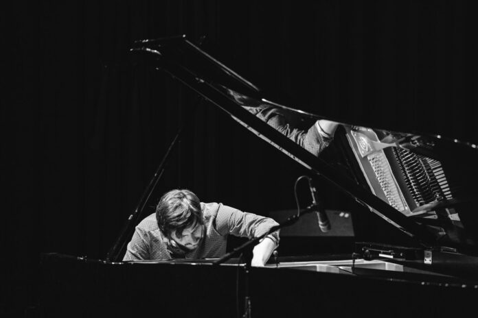 Schwarzweißfoto eines Pianisten am geöffneten Konzertflügel. Der Musiker beugt sich konzentriert über die Klaviersaiten und greift mit einer Hand in den Flügel, während ein Mikrofon nahe der Saiten positioniert ist. Das Bühnenlicht hebt Hände, Saiten und Mechanik hervor, der Hintergrund bleibt dunkel.