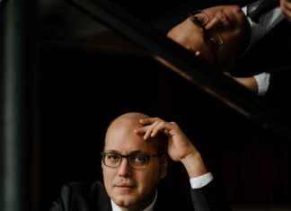 Gottlieb Wallisch bei “Ewige Hoffnung” / Kulturforum Prag Das Porträt zeigt einen aufgeklappten Klavierflügel, hinter dem ein mittelalter weißer Mann mit Brille in Anzug und Krawatte sitzt. Ein Arm liegt auf dem Klavier auf, mit dem anderen stützt er seinen Kopf ab. Oben im offenen Klavierflügel spiegelt sich das Innere des Instrumentes sowie der Kopf des Mannes.