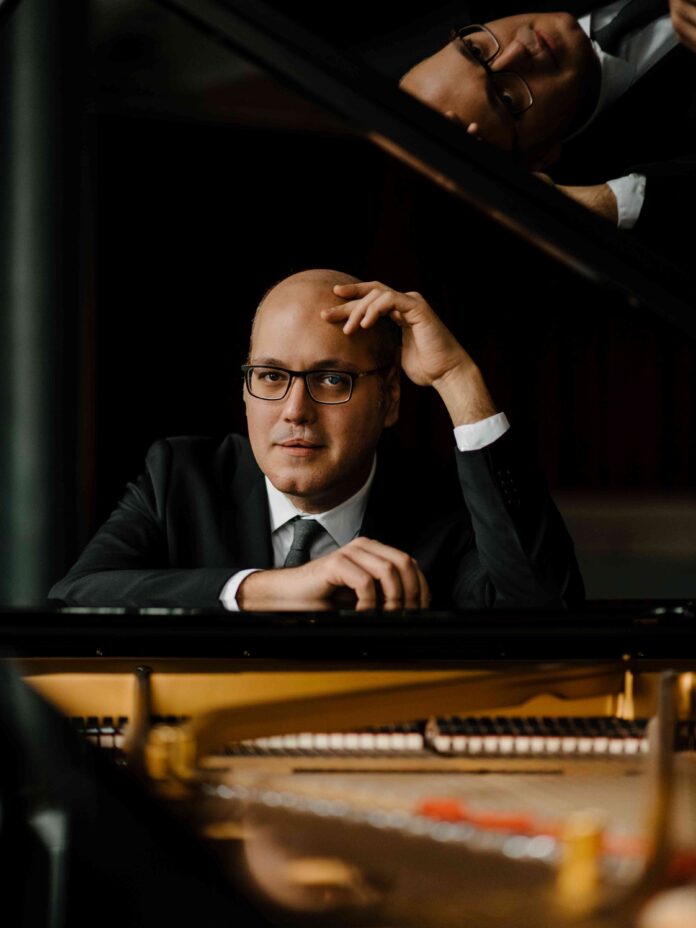 Das Porträt zeigt einen aufgeklappten Klavierflügel, hinter dem ein mittelalter weißer Mann mit Brille in Anzug und Krawatte sitzt. Ein Arm liegt auf dem Klavier auf, mit dem anderen stützt er seinen Kopf ab. Oben im offenen Klavierflügel spiegelt sich das Innere des Instrumentes sowie der Kopf des Mannes.