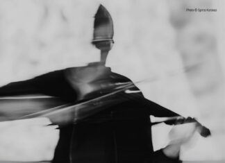 Mia Zabelka / Botschaft Oslo Das Bild ist ein schwarz-weißes Konzertplakat im Hochformat. Im Hintergrund ist eine abstrakte, leicht verschwommene Fotografie einer Person zu sehen, die eine Violine spielt. Die Bewegung des Arms und des Bogens ist verwischt, wodurch ein Eindruck von Dynamik, Klang und körperlicher Präsenz entsteht. Das Gesicht ist nicht klar erkennbar, was dem Bild einen anonymen, künstlerischen Charakter verleiht. Die Gesamtwirkung ist expressiv und atmosphärisch. Die Farbgestaltung ist reduziert auf Schwarz-, Grau- und Weißtöne. Der untere Bereich des Plakats ist dunkler gehalten, um den hell gesetzten Text gut lesbar hervorzuheben.