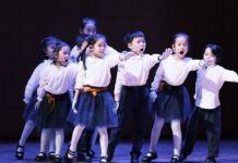 Die Zauberflöte von Kindern für Kinder / Konzertabend / Kulturforum Peking Das Bild zeigt eine Gruppe von Kindern auf einer beleuchteten Bühne während einer Opernaufführung. Die Kinder tragen weiße Oberteile, dunkle Röcke bzw. Hosen sowie weiße Handschuhe und singen, während sie synchron eine Armbewegung nach vorne ausführen. Sie stehen in mehreren Reihen und wirken konzentriert und engagiert. Die dunkle Bühnenkulisse hebt die Kinder deutlich hervor und unterstreicht den Aufführungscharakter der Szene.