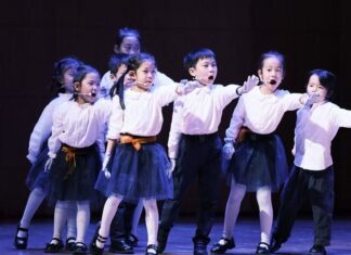 Die Zauberflöte von Kindern für Kinder / Konzertabend / Kulturforum Peking Das Bild zeigt eine Gruppe von Kindern auf einer beleuchteten Bühne während einer Opernaufführung. Die Kinder tragen weiße Oberteile, dunkle Röcke bzw. Hosen sowie weiße Handschuhe und singen, während sie synchron eine Armbewegung nach vorne ausführen. Sie stehen in mehreren Reihen und wirken konzentriert und engagiert. Die dunkle Bühnenkulisse hebt die Kinder deutlich hervor und unterstreicht den Aufführungscharakter der Szene.