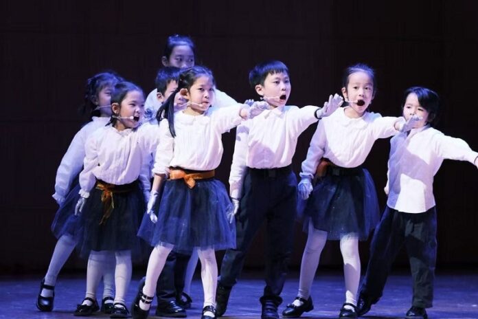 altKFPeking Das Bild zeigt eine Gruppe von Kindern auf einer beleuchteten Bühne während einer Opernaufführung. Die Kinder tragen weiße Oberteile, dunkle Röcke bzw. Hosen sowie weiße Handschuhe und singen, während sie synchron eine Armbewegung nach vorne ausführen. Sie stehen in mehreren Reihen und wirken konzentriert und engagiert. Die dunkle Bühnenkulisse hebt die Kinder deutlich hervor und unterstreicht den Aufführungscharakter der Szene.