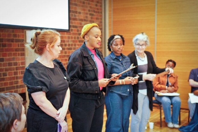 Bild: Catherine Boyd Auf dem Bild sieht man vier Personen auf einer Bühne, die miteinander etwas vortragen.