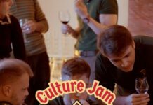 Culture Jam: Myths & Magic / Kulturforum Warschau Mehrere junge Erwachsene sitzen und stehen um einen großen Tisch und arbeiten gemeinsam an Laptops. Einige schauen auf die Bildschirme, andere beugen sich über die Arbeitsplätze und diskutieren. Die Szene vermittelt eine konzentrierte, lebendige Arbeitsatmosphäre bei einem gemeinsamen Projekt. In der Mitte des Bildes ist ein großes grafisches Logo eingeblendet: ein Wappen im Stil eines mittelalterlichen Schildes, in den Farben der österreichischen Flagge Rot-Weiß-Rot, mit einem stilisierten grünen Drachen in der Mitte. Über und unter dem Wappen steht der Schriftzug „Culture Jam – Myths & Magic“.