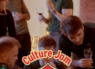Culture Jam: Myths & Magic / Kulturforum Warschau Mehrere junge Erwachsene sitzen und stehen um einen großen Tisch und arbeiten gemeinsam an Laptops. Einige schauen auf die Bildschirme, andere beugen sich über die Arbeitsplätze und diskutieren. Die Szene vermittelt eine konzentrierte, lebendige Arbeitsatmosphäre bei einem gemeinsamen Projekt. In der Mitte des Bildes ist ein großes grafisches Logo eingeblendet: ein Wappen im Stil eines mittelalterlichen Schildes, in den Farben der österreichischen Flagge Rot-Weiß-Rot, mit einem stilisierten grünen Drachen in der Mitte. Über und unter dem Wappen steht der Schriftzug „Culture Jam – Myths & Magic“.