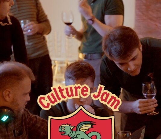 Culture Jam: Myths & Magic / Kulturforum Warschau Mehrere junge Erwachsene sitzen und stehen um einen großen Tisch und arbeiten gemeinsam an Laptops. Einige schauen auf die Bildschirme, andere beugen sich über die Arbeitsplätze und diskutieren. Die Szene vermittelt eine konzentrierte, lebendige Arbeitsatmosphäre bei einem gemeinsamen Projekt. In der Mitte des Bildes ist ein großes grafisches Logo eingeblendet: ein Wappen im Stil eines mittelalterlichen Schildes, in den Farben der österreichischen Flagge Rot-Weiß-Rot, mit einem stilisierten grünen Drachen in der Mitte. Über und unter dem Wappen steht der Schriftzug „Culture Jam – Myths & Magic“.
