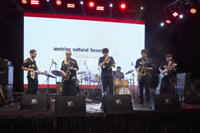 Es ist die Band Shake Stew auf der Bühne zu sehen. Im Hintergrund das Logo des Kulturforums