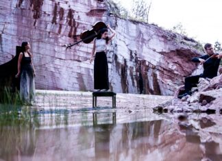 accio piano trio / Kulturforum Berlin Das Bild zeigt das accio piano trio in einer eindrucksvollen Naturkulisse mit Felswand und Wasser im Vordergrund. In der Mitte steht die Cellistin mit ihrem Instrument, links befindet sich die Pianistin am Flügel und rechts spielt der Geiger Clemens Böck auf einem Felsen. Die drei tragen elegante, dunkle Konzertkleidung. Die atmosphärische Inszenierung verbindet Natur und klassische Musik auf poetische Weise.