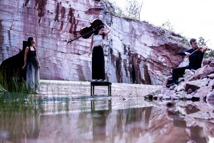 Das Bild zeigt das accio piano trio in einer eindrucksvollen Naturkulisse mit Felswand und Wasser im Vordergrund. In der Mitte steht die Cellistin mit ihrem Instrument, links befindet sich die Pianistin am Flügel und rechts spielt der Geiger Clemens Böck auf einem Felsen. Die drei tragen elegante, dunkle Konzertkleidung. Die atmosphärische Inszenierung verbindet Natur und klassische Musik auf poetische Weise.
