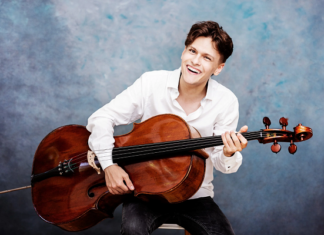 Konzert „Transformation“ / Kulturforum Istanbul & Botschaft Ankara Das Bild zeigt einen lächelnden jungen Mann in weißem Hemd, der im Sitzen ein Cello hält und vor einem blau-grauen Studiohintergrund musiziert.