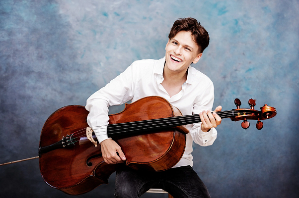 Das Bild zeigt einen lächelnden jungen Mann in weißem Hemd, der im Sitzen ein Cello hält und vor einem blau-grauen Studiohintergrund musiziert.