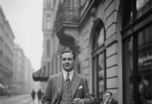 Gershwin in Vienna / Kulturforum New York Das Bild zeigt einen elegant gekleideten Mann in den 1920er-Jahren, der auf einem Gehsteig in einer europäischen Stadt steht. Er trägt einen dreiteiligen Anzug mit Krawatte und Einstecktuch und hält eine Zigarette in der Hand. Im Hintergrund sind Straßencafés mit sitzenden Gästen, kleine runde Tische mit Kaffeetassen sowie mehrstöckige Gebäude mit Balkonen zu sehen. Die Szene ist in Schwarz-Weiß gehalten und vermittelt eine ruhige, urbane Atmosphäre der frühen Moderne.