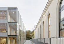 Dietmar Feichtinger / Kulturforum New York Modernes Schulgebäude mit einem schmalen, offenen Verbindungsgang zwischen einem historischen Bau mit Rundbogenfenstern (rechts) und einem zeitgenössischen Glas- und Metallbau (links). Der Gang wird von einem Metallgeländer begleitet und führt über eine Brücke. Durch große Fenster ist im unteren Bereich ein heller Innenraum mit Holzboden und mehreren Tischen sichtbar. Im Hintergrund sind Bäume zu sehen.