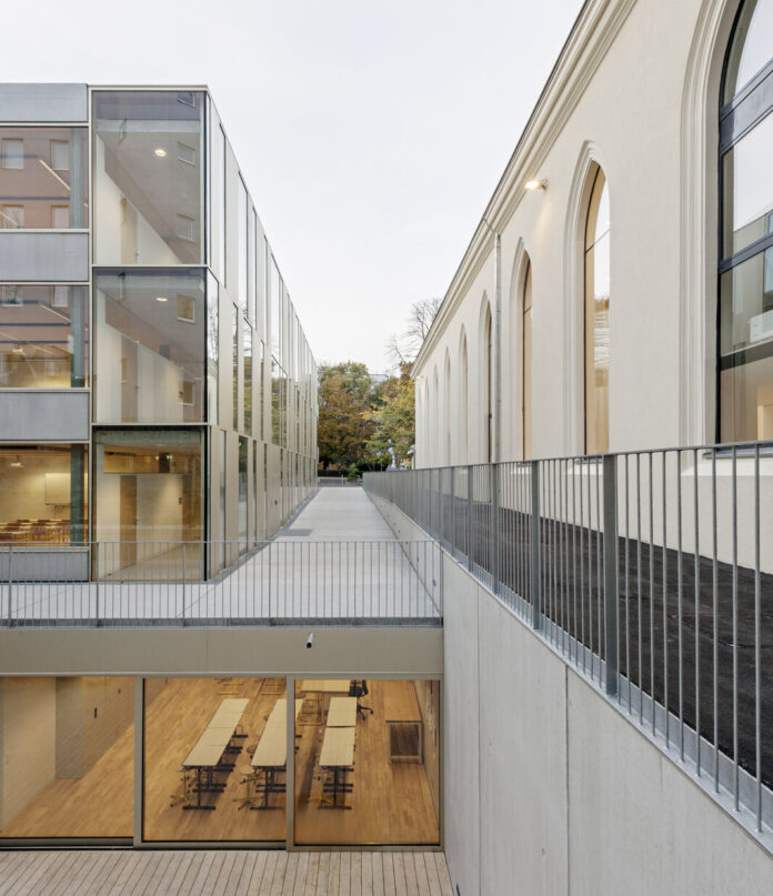 Bild: DIETMAR FEICHTINGER ARCHITECTES, School Extension in Vienna, 2010, Image: Hertha Hurnaus Modernes Schulgebäude mit einem schmalen, offenen Verbindungsgang zwischen einem historischen Bau mit Rundbogenfenstern (rechts) und einem zeitgenössischen Glas- und Metallbau (links). Der Gang wird von einem Metallgeländer begleitet und führt über eine Brücke. Durch große Fenster ist im unteren Bereich ein heller Innenraum mit Holzboden und mehreren Tischen sichtbar. Im Hintergrund sind Bäume zu sehen.