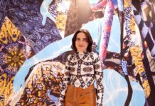 Sunny Pfalzer – Cripto Sirenas / Kulturforum Ottawa Das Bild zeigt die österreichische Künstlerin Sunny Pfalzer, die vor einem ihrer Kunstwerke steht und lächelt.