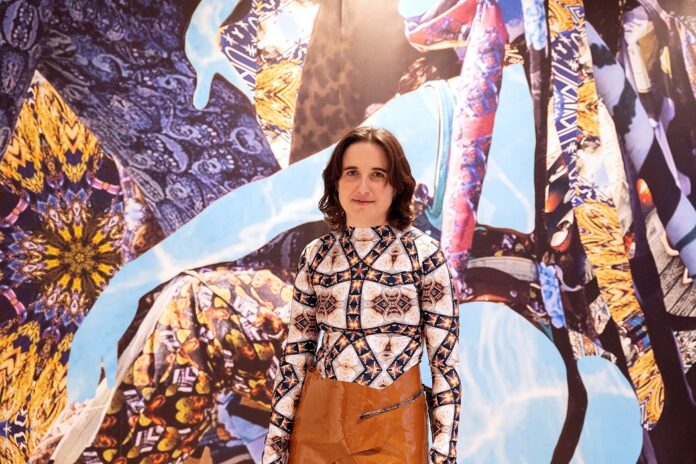 Das Bild zeigt die österreichische Künstlerin Sunny Pfalzer, die vor einem ihrer Kunstwerke steht und lächelt.
