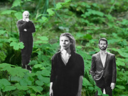 Da kommt noch wer / Botschaft Oslo In einem dichten, grünen Wald mit Farnen und Unterholz stehen drei schwarz-weiße, ausgeschnittene Personenfiguren, die wie in die Natur hineingestellt wirken. Im Vordergrund befindet sich eine Frau mit schulterlangem Haar und dunkler Kleidung, die geradeaus blickt. Etwas dahinter stehen zwei Männer: links eine Person mit nachdenklicher Haltung und angewinkeltem Arm, rechts ein Mann in Anzugjacke mit offenem Hemd, der frontal nach vorn schaut. Der starke Kontrast zwischen den grauen Figuren und der lebendigen, grünen Waldumgebung erzeugt einen collageartigen, surrealen Eindruck.