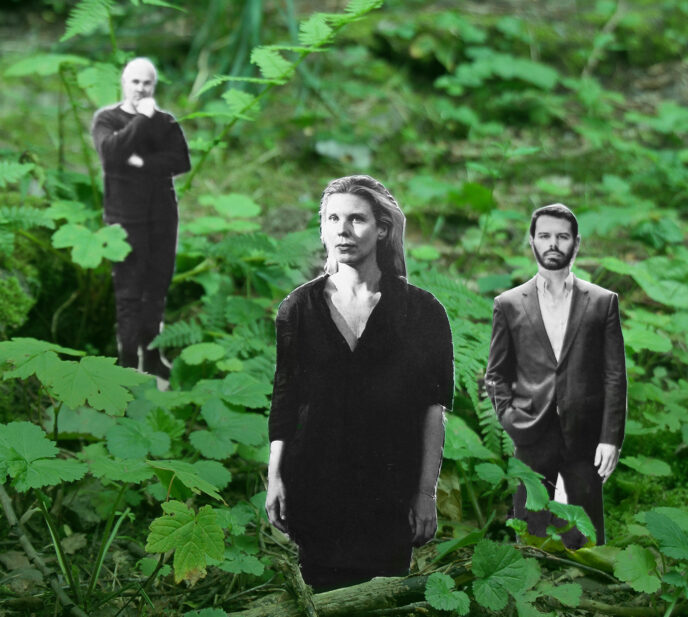 In einem dichten, grünen Wald mit Farnen und Unterholz stehen drei schwarz-weiße, ausgeschnittene Personenfiguren, die wie in die Natur hineingestellt wirken. Im Vordergrund befindet sich eine Frau mit schulterlangem Haar und dunkler Kleidung, die geradeaus blickt. Etwas dahinter stehen zwei Männer: links eine Person mit nachdenklicher Haltung und angewinkeltem Arm, rechts ein Mann in Anzugjacke mit offenem Hemd, der frontal nach vorn schaut. Der starke Kontrast zwischen den grauen Figuren und der lebendigen, grünen Waldumgebung erzeugt einen collageartigen, surrealen Eindruck.