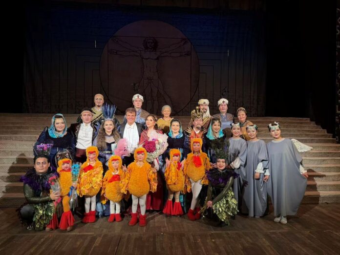 Gruppenfoto auf einer Theaterbühne nach einer Opernaufführung. In der Mitte steht der Dirigent Matthias Fletzberger in Frack, umgeben von Sängerinnen, Sängern und Darstellern in aufwendigen, farbenfrohen Kostümen, darunter auch mehrere Kinder in vogelähnlichen Bühnenoutfits. Die Mitwirkenden halten Blumen und blicken lächelnd in die Kamera..