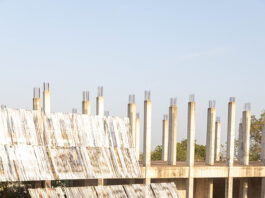 Ausstellung: Limbo Engawa / Kulturforum Accra Das Bild zeigt eine Gruppe von Menschen, die im Freien auf dem Boden sitzt und einer Präsentation zuhört, die vor einem teilweise fertiggestellten Betongebäude mit vielen freistehenden Säulen stattfindet. Im Vordergrund sind geneigte, reflektierende Metallrahmen, die mit Bannerstreifen bezogen sind, aufgebaut, die als eine Art temporäre Bühne oder Installation dienen. Die Umgebung wirkt ländlich, mit roter Erde, vereinzelten Bäumen und warmem Sonnenlicht.