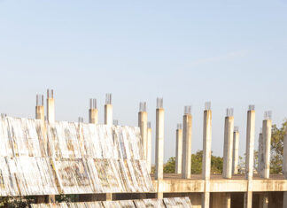 Ausstellung: Limbo Engawa / Kulturforum Accra Das Bild zeigt eine Gruppe von Menschen, die im Freien auf dem Boden sitzt und einer Präsentation zuhört, die vor einem teilweise fertiggestellten Betongebäude mit vielen freistehenden Säulen stattfindet. Im Vordergrund sind geneigte, reflektierende Metallrahmen, die mit Bannerstreifen bezogen sind, aufgebaut, die als eine Art temporäre Bühne oder Installation dienen. Die Umgebung wirkt ländlich, mit roter Erde, vereinzelten Bäumen und warmem Sonnenlicht.