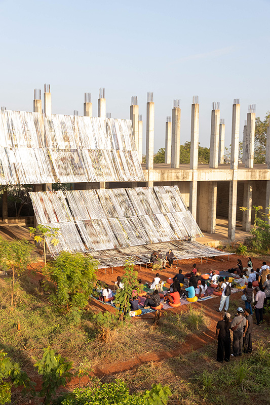 Das Bild zeigt eine Gruppe von Menschen, die im Freien auf dem Boden sitzt und einer Präsentation zuhört, die vor einem teilweise fertiggestellten Betongebäude mit vielen freistehenden Säulen stattfindet. Im Vordergrund sind geneigte, reflektierende Metallrahmen, die mit Bannerstreifen bezogen sind, aufgebaut, die als eine Art temporäre Bühne oder Installation dienen. Die Umgebung wirkt ländlich, mit roter Erde, vereinzelten Bäumen und warmem Sonnenlicht.