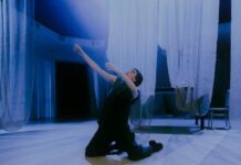 HiStorieS – Angelus Novus / Kulturforum Berlin Das Bild zeigt die Performerin Loulou Omer. Sie kniet in der Mitte des Raumes auf dem Boden. Sie trägt schwarze Kleidung und ihr Blick geht nach links oben zum Licht. Von der Decke hängen weiße, durchsichtige Tücher, die den hellen Tanzboden fast berühren. Im Hintergrund stehen ein Tisch und ein Stuhl, die von den weißen Tüchern bedeckt sind. Das von oben einfallende Licht betont die engelsgleiche Atmosphäre