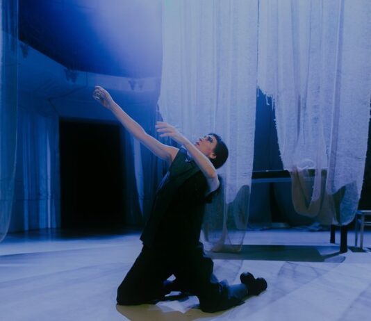 HiStorieS – Angelus Novus / Kulturforum Berlin Das Bild zeigt die Performerin Loulou Omer. Sie kniet in der Mitte des Raumes auf dem Boden. Sie trägt schwarze Kleidung und ihr Blick geht nach links oben zum Licht. Von der Decke hängen weiße, durchsichtige Tücher, die den hellen Tanzboden fast berühren. Im Hintergrund stehen ein Tisch und ein Stuhl, die von den weißen Tüchern bedeckt sind. Das von oben einfallende Licht betont die engelsgleiche Atmosphäre