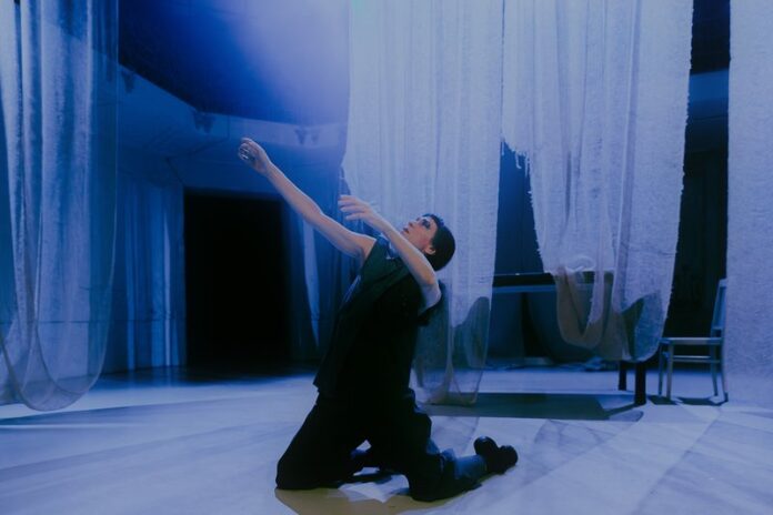 Das Bild zeigt die Performerin Loulou Omer. Sie kniet in der Mitte des Raumes auf dem Boden. Sie trägt schwarze Kleidung und ihr Blick geht nach links oben zum Licht. Von der Decke hängen weiße, durchsichtige Tücher, die den hellen Tanzboden fast berühren. Im Hintergrund stehen ein Tisch und ein Stuhl, die von den weißen Tüchern bedeckt sind. Das von oben einfallende Licht betont die engelsgleiche Atmosphäre