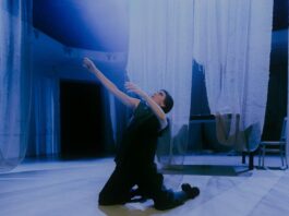 HiStorieS – Angelus Novus / Kulturforum Berlin Das Bild zeigt die Performerin Loulou Omer. Sie kniet in der Mitte des Raumes auf dem Boden. Sie trägt schwarze Kleidung und ihr Blick geht nach links oben zum Licht. Von der Decke hängen weiße, durchsichtige Tücher, die den hellen Tanzboden fast berühren. Im Hintergrund stehen ein Tisch und ein Stuhl, die von den weißen Tüchern bedeckt sind. Das von oben einfallende Licht betont die engelsgleiche Atmosphäre.