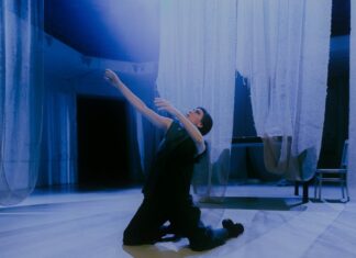 HiStorieS – Angelus Novus / Kulturforum Berlin Das Bild zeigt die Performerin Loulou Omer. Sie kniet in der Mitte des Raumes auf dem Boden. Sie trägt schwarze Kleidung und ihr Blick geht nach links oben zum Licht. Von der Decke hängen weiße, durchsichtige Tücher, die den hellen Tanzboden fast berühren. Im Hintergrund stehen ein Tisch und ein Stuhl, die von den weißen Tüchern bedeckt sind. Das von oben einfallende Licht betont die engelsgleiche Atmosphäre.