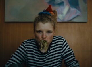 Film von Florian Pochlatko / Kulturforum Prag Das Bild zeigt eine junge Frau mit vollem Mund voller Spaghetti, die an einem Esstisch sitzt und in die Kamera starrt.