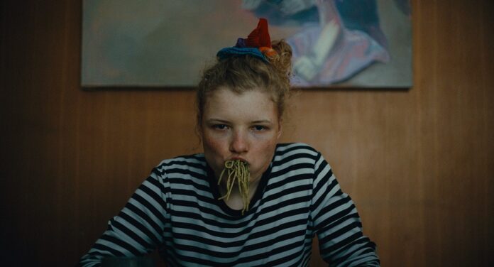 Das Bild zeigt eine junge Frau mit vollem Mund voller Spaghetti, die an einem Esstisch sitzt und in die Kamera starrt.