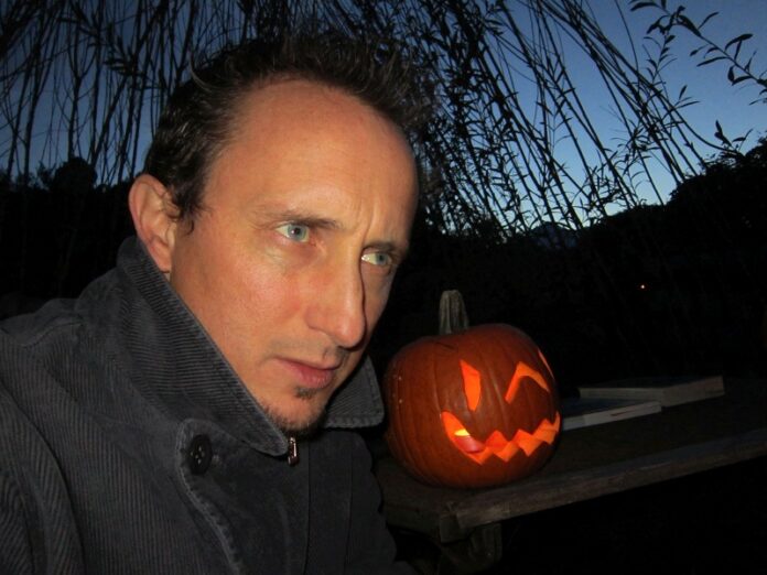 Das Bild zeigt Christian Kössler, der draußen in der Dämmerung neben einem leuchtenden Halloween-Kürbis sitzt.