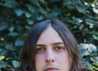 Elias Hirschl / Podcast wissensArt Ein Portrait des Künstlers Elias Hirschl. Er trägt eine schwarze Lederjacke und längeres Haar und blickt direkt in die Kamera. Im Hintergrund grüne Blätter.