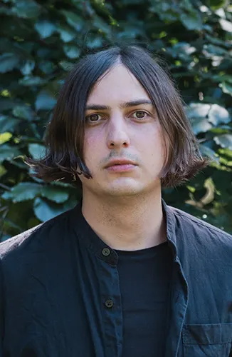 Ein Portrait des Künstlers Elias Hirschl. Er trägt eine schwarze Lederjacke und längeres Haar und blickt direkt in die Kamera. Im Hintergrund grüne Blätter.