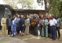 Strategic Roundtable / Eröffnung Kulturforum Accra Das Bild zeigt ein abendliches Live-Konzert auf einer beleuchteten Bühne im Freien. Auf der Bühne stehen mehrere Musikerinnen und Musiker und spielen verschiedene Instrumente, darunter Gitarre, Saxofon und Schlagzeug. Über ihnen befindet sich ein Metallgerüst mit bunten Bühnenlichtern in Blau, Lila und Gelb. Im Hintergrund der Band ist eine große LED-Leinwand mit einem abstrakten, farbintensiven Muster in Orange-, Pink- und Gelbtönen zu sehen. Im Vordergrund sitzt ein Publikum an Tischen und schaut in Richtung der Bühne. Die Personen sind größtenteils nur als dunkle Silhouetten erkennbar. Die Atmosphäre wirkt lebendig und festlich, mit stimmungsvoller Beleuchtung und Konzertambiente unter freiem Himmel.