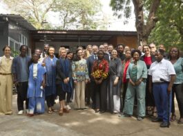 Strategic Roundtable / Eröffnung Kulturforum Accra Das Bild zeigt ein abendliches Live-Konzert auf einer beleuchteten Bühne im Freien. Auf der Bühne stehen mehrere Musikerinnen und Musiker und spielen verschiedene Instrumente, darunter Gitarre, Saxofon und Schlagzeug. Über ihnen befindet sich ein Metallgerüst mit bunten Bühnenlichtern in Blau, Lila und Gelb. Im Hintergrund der Band ist eine große LED-Leinwand mit einem abstrakten, farbintensiven Muster in Orange-, Pink- und Gelbtönen zu sehen. Im Vordergrund sitzt ein Publikum an Tischen und schaut in Richtung der Bühne. Die Personen sind größtenteils nur als dunkle Silhouetten erkennbar. Die Atmosphäre wirkt lebendig und festlich, mit stimmungsvoller Beleuchtung und Konzertambiente unter freiem Himmel.