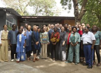 Strategic Roundtable / Eröffnung Kulturforum Accra Das Bild zeigt ein abendliches Live-Konzert auf einer beleuchteten Bühne im Freien. Auf der Bühne stehen mehrere Musikerinnen und Musiker und spielen verschiedene Instrumente, darunter Gitarre, Saxofon und Schlagzeug. Über ihnen befindet sich ein Metallgerüst mit bunten Bühnenlichtern in Blau, Lila und Gelb. Im Hintergrund der Band ist eine große LED-Leinwand mit einem abstrakten, farbintensiven Muster in Orange-, Pink- und Gelbtönen zu sehen. Im Vordergrund sitzt ein Publikum an Tischen und schaut in Richtung der Bühne. Die Personen sind größtenteils nur als dunkle Silhouetten erkennbar. Die Atmosphäre wirkt lebendig und festlich, mit stimmungsvoller Beleuchtung und Konzertambiente unter freiem Himmel.