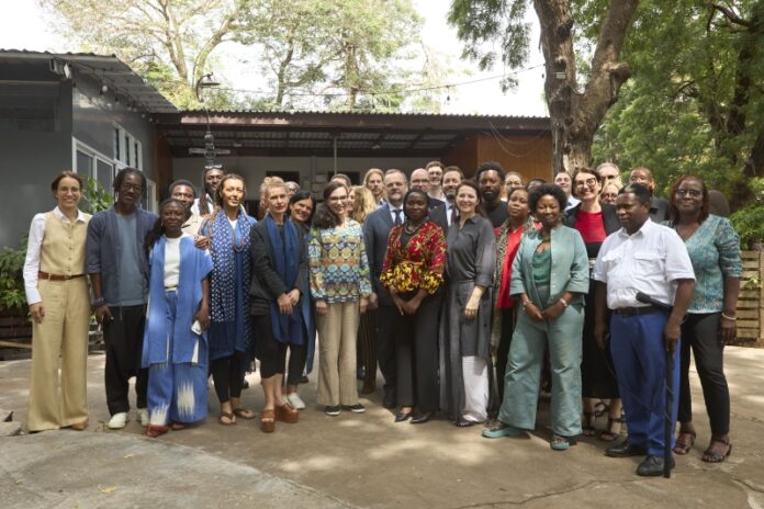 Das Bild zeigt ein abendliches Live-Konzert auf einer beleuchteten Bühne im Freien. Auf der Bühne stehen mehrere Musikerinnen und Musiker und spielen verschiedene Instrumente, darunter Gitarre, Saxofon und Schlagzeug. Über ihnen befindet sich ein Metallgerüst mit bunten Bühnenlichtern in Blau, Lila und Gelb. Im Hintergrund der Band ist eine große LED-Leinwand mit einem abstrakten, farbintensiven Muster in Orange-, Pink- und Gelbtönen zu sehen. Im Vordergrund sitzt ein Publikum an Tischen und schaut in Richtung der Bühne. Die Personen sind größtenteils nur als dunkle Silhouetten erkennbar. Die Atmosphäre wirkt lebendig und festlich, mit stimmungsvoller Beleuchtung und Konzertambiente unter freiem Himmel.