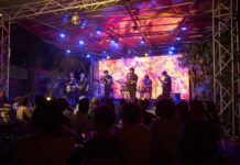 Konzert von Shake Stew / Eröffnung Kulturforum Accra Das Bild zeigt eine große Gruppe von etwa 20 erwachsenen Personen, die im Freien für ein Gruppenfoto posieren. Sie stehen dicht nebeneinander auf einem betonierten Hof unter schattenspendenden Bäumen. Im Hintergrund ist ein einstöckiges Gebäude mit Wellblechdach und Holzelementen zu sehen. Die Gruppe ist vielfältig in Bezug auf Alter, Geschlecht und Hautfarbe. Die Personen tragen unterschiedliche Kleidung, von Anzügen und eleganten Kleidern bis hin zu farbenfrohen, gemusterten Outfits und legerer Alltagskleidung. Einige lächeln direkt in die Kamera, andere blicken leicht zur Seite. Die Stimmung wirkt zugleich freundlich und formell. Natürliches Tageslicht fällt durch die Bäume und sorgt für eine helle, warme Atmosphäre.