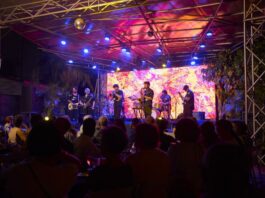 Konzert von Shake Stew / Eröffnung Kulturforum Accra Das Bild zeigt eine große Gruppe von etwa 20 erwachsenen Personen, die im Freien für ein Gruppenfoto posieren. Sie stehen dicht nebeneinander auf einem betonierten Hof unter schattenspendenden Bäumen. Im Hintergrund ist ein einstöckiges Gebäude mit Wellblechdach und Holzelementen zu sehen. Die Gruppe ist vielfältig in Bezug auf Alter, Geschlecht und Hautfarbe. Die Personen tragen unterschiedliche Kleidung, von Anzügen und eleganten Kleidern bis hin zu farbenfrohen, gemusterten Outfits und legerer Alltagskleidung. Einige lächeln direkt in die Kamera, andere blicken leicht zur Seite. Die Stimmung wirkt zugleich freundlich und formell. Natürliches Tageslicht fällt durch die Bäume und sorgt für eine helle, warme Atmosphäre.