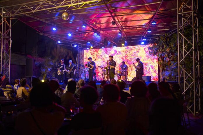 Das Bild zeigt eine große Gruppe von etwa 20 erwachsenen Personen, die im Freien für ein Gruppenfoto posieren. Sie stehen dicht nebeneinander auf einem betonierten Hof unter schattenspendenden Bäumen. Im Hintergrund ist ein einstöckiges Gebäude mit Wellblechdach und Holzelementen zu sehen. Die Gruppe ist vielfältig in Bezug auf Alter, Geschlecht und Hautfarbe. Die Personen tragen unterschiedliche Kleidung, von Anzügen und eleganten Kleidern bis hin zu farbenfrohen, gemusterten Outfits und legerer Alltagskleidung. Einige lächeln direkt in die Kamera, andere blicken leicht zur Seite. Die Stimmung wirkt zugleich freundlich und formell. Natürliches Tageslicht fällt durch die Bäume und sorgt für eine helle, warme Atmosphäre.