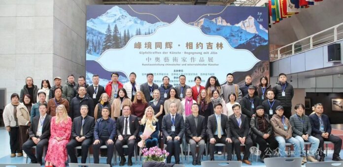 Bild: Changchun Foreign Affairs Office Das Bild zeigt ein Gruppenfoto der beteiligten Künstlerinnen, Künstler und Gäste bei der Ausstellung „Gipfeltreffen der Künste – Begegnung mit Jilin“. Die Gruppe ist vor dem Ausstellungsplakat mit Bergmotiv und chinesischer Schrift auf einer Bühne versammelt.