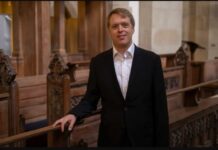 Donner in der Domkirche / Botschaft Oslo Stefan Donner steht in einem Innenraum einer Kirche. Er hat kurze blonde Haare, trägt einen schwarzen Anzug mit weißem Hemd und lächelt leicht in die Kamera. Mit einer Hand stützt er sich locker auf eine kunstvoll geschnitzte Holzbank. Im Hintergrund sind weitere hölzerne Sitzreihen, Säulen und warme Beleuchtung zu sehen.