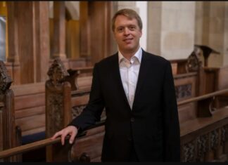 Donner in der Domkirche / Botschaft Oslo Stefan Donner steht in einem Innenraum einer Kirche. Er hat kurze blonde Haare, trägt einen schwarzen Anzug mit weißem Hemd und lächelt leicht in die Kamera. Mit einer Hand stützt er sich locker auf eine kunstvoll geschnitzte Holzbank. Im Hintergrund sind weitere hölzerne Sitzreihen, Säulen und warme Beleuchtung zu sehen.